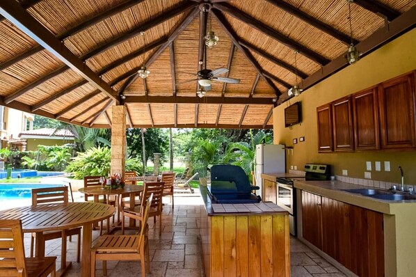 Relax in a shaded outdoor space with a dining set, barbecue grill, and kitchen area surrounded by lush greenery and a nearby pool.