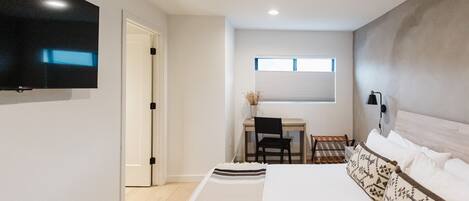 A bedroom with a bed, a desk, a chair, a TV, and a window.