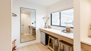A modern kitchen with a large mirror, wooden stools, a small refrigerator, and a window with a view of a vehicle outside.