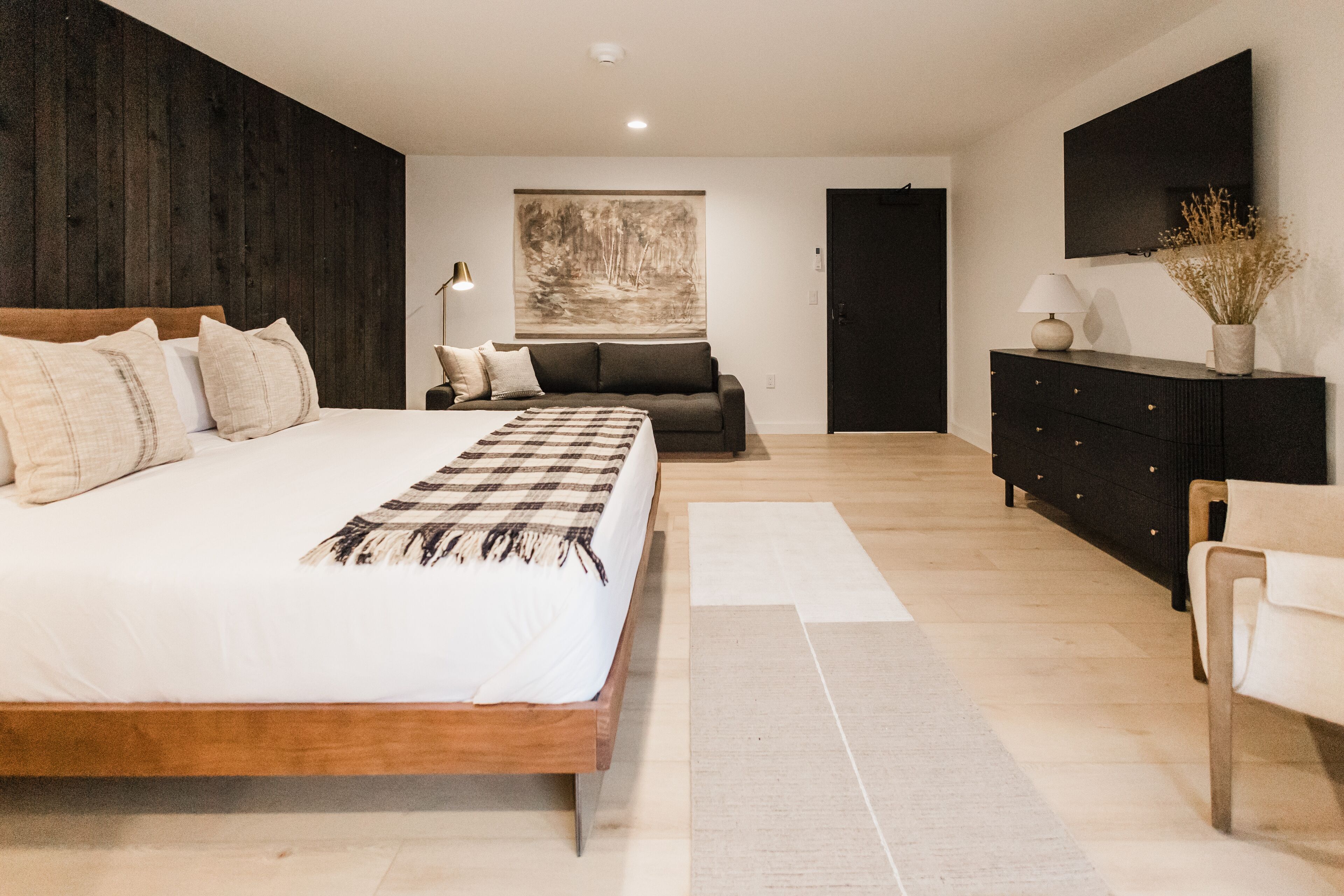 A modern bedroom with a large bed, a dark wooden accent wall, a gray sofa, a black dresser, and a painting on the wall.