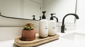 Une salle de bain avec un lavabo blanc, deux flacons de lotion, une petite plante en pot et un plateau en bois.