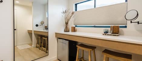 A modern kitchen with a large mirror, wooden stools, a built-in refrigerator, and a window with blinds.