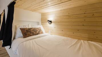 A neatly made bed with white linens and a patterned pillow, a wooden ceiling, and a wall-mounted light fixture.