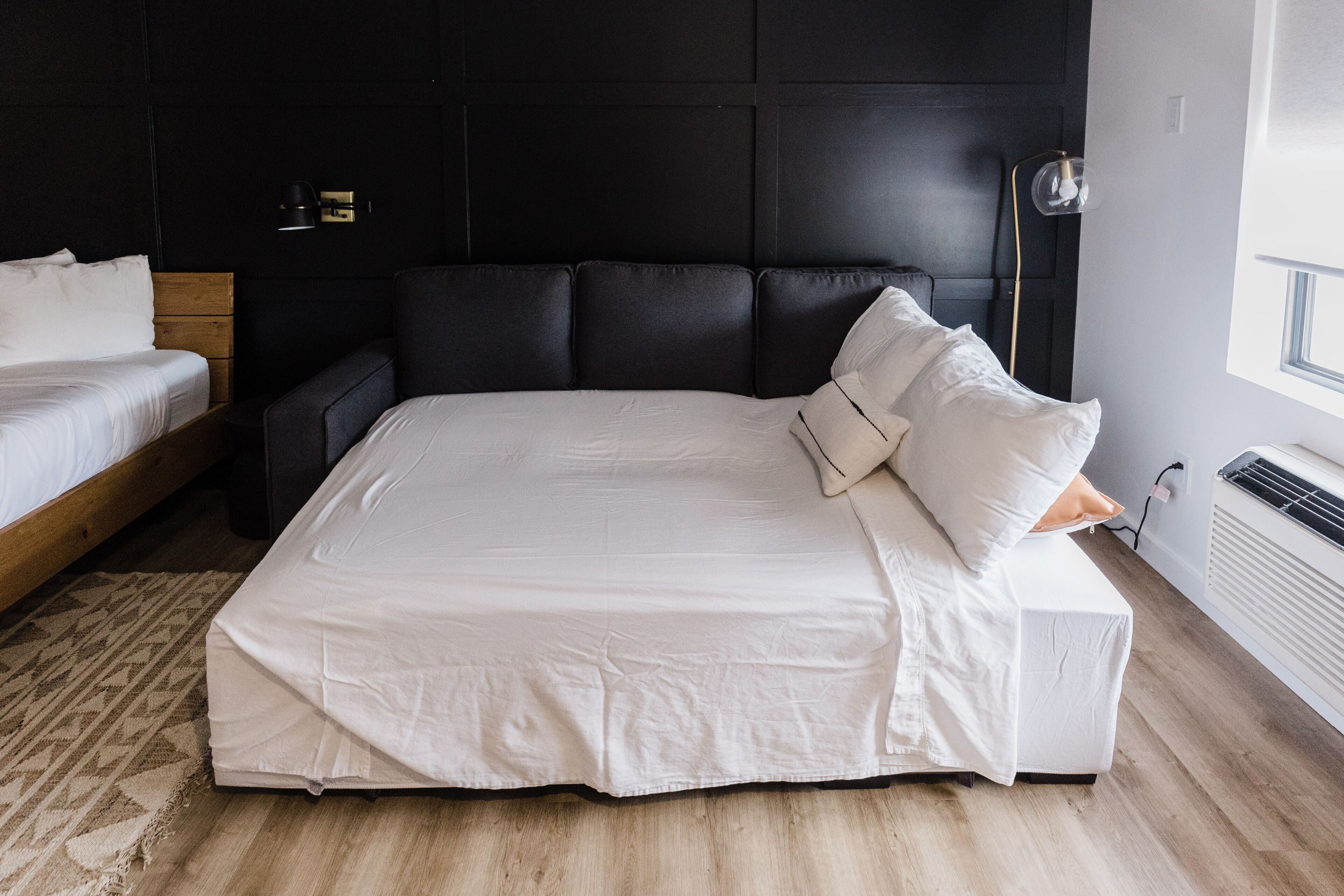 A modern bedroom with a large bed, a smaller bed, a dark headboard, a bedside table, and a lamp.