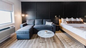 Une chambre d’hôtel moderne avec un canapé d’angle, un lit simple, une table basse ronde et une grande fenêtre.