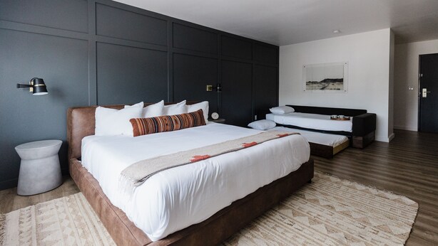 Un dormitorio moderno con una cama grande, una cama más pequeña y un banco. Hay dos mesitas de noche y una lámpara de pared.