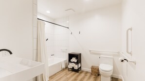 A bathroom with a white sink, bathtub, and toilet. There is a shelf with towels and a basket on the floor. - Wonderstone Single Queen ADA Guest Room (Columbia Falls)