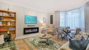 Ein Wohnzimmer mit Kamin, Fernseher, Bücherregal, Essbereich mit Tisch und Stühlen sowie einem großen Fenster mit Vorhängen.