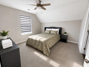 Une chambre à coucher comprenant un lit, une table de chevet, un ventilateur de plafond, une fenêtre avec des stores et une porte.