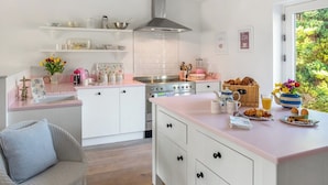 Une cuisine moderne avec des armoires blanches, un plan de travail rose et un îlot central. On y trouve une cuisinière, un évier et divers ustensiles de cuisine. Une chaise en osier est visible au premier plan.