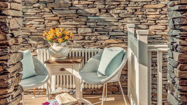 Un intérieur avec un mur en pierre, deux fauteuils en osier, une petite table sur laquelle est posé un vase de fleurs et un radiateur.
