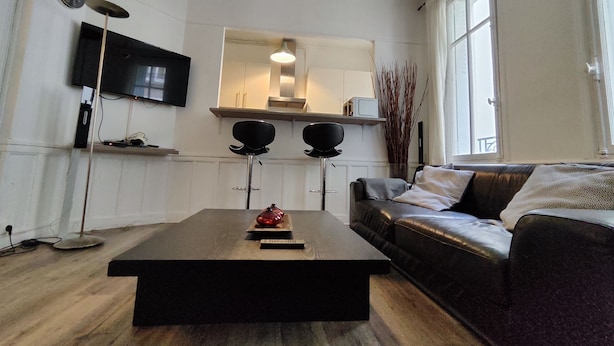 Ein modernes Wohnzimmer mit einem Holztisch, einem schwarzen Sofa, einem Flachbildfernseher und einem großen Spiegel über einem weißen Schrank.