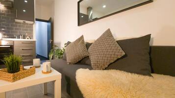 A modern living room with a sofa, two patterned pillows, a fluffy throw, a coffee table with a plant and mugs, and a kitchen area with a tiled backsplash and a stove.