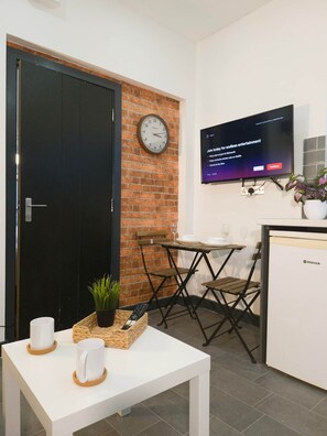 A room with a black door, a clock on the wall, a television displaying an advertisement, a small table with a plant and two cups, and a refrigerator.