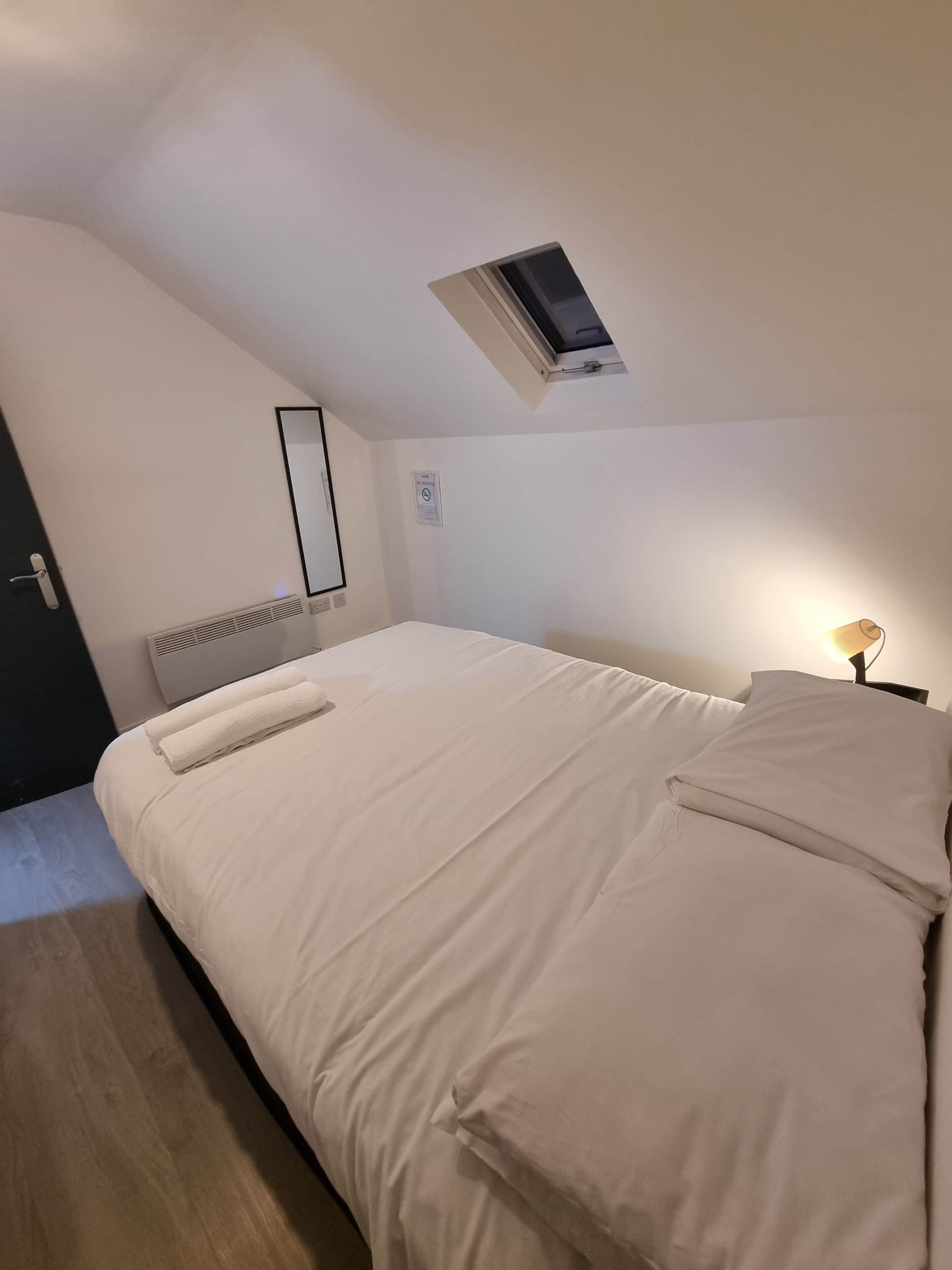 Een slaapkamer met een groot bed, een klein raam, een radiator, een deur en een nachtlampje.