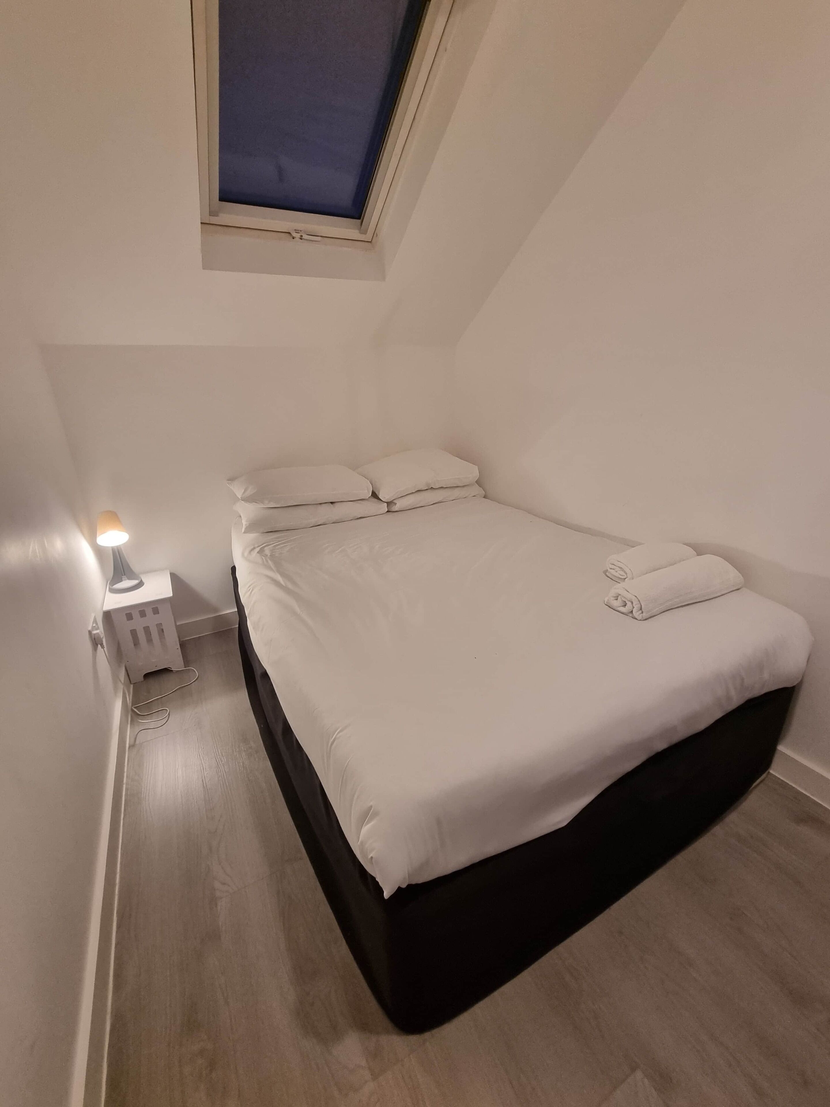 A small, neatly arranged bedroom with a single bed, a nightstand, a lamp, and a window.