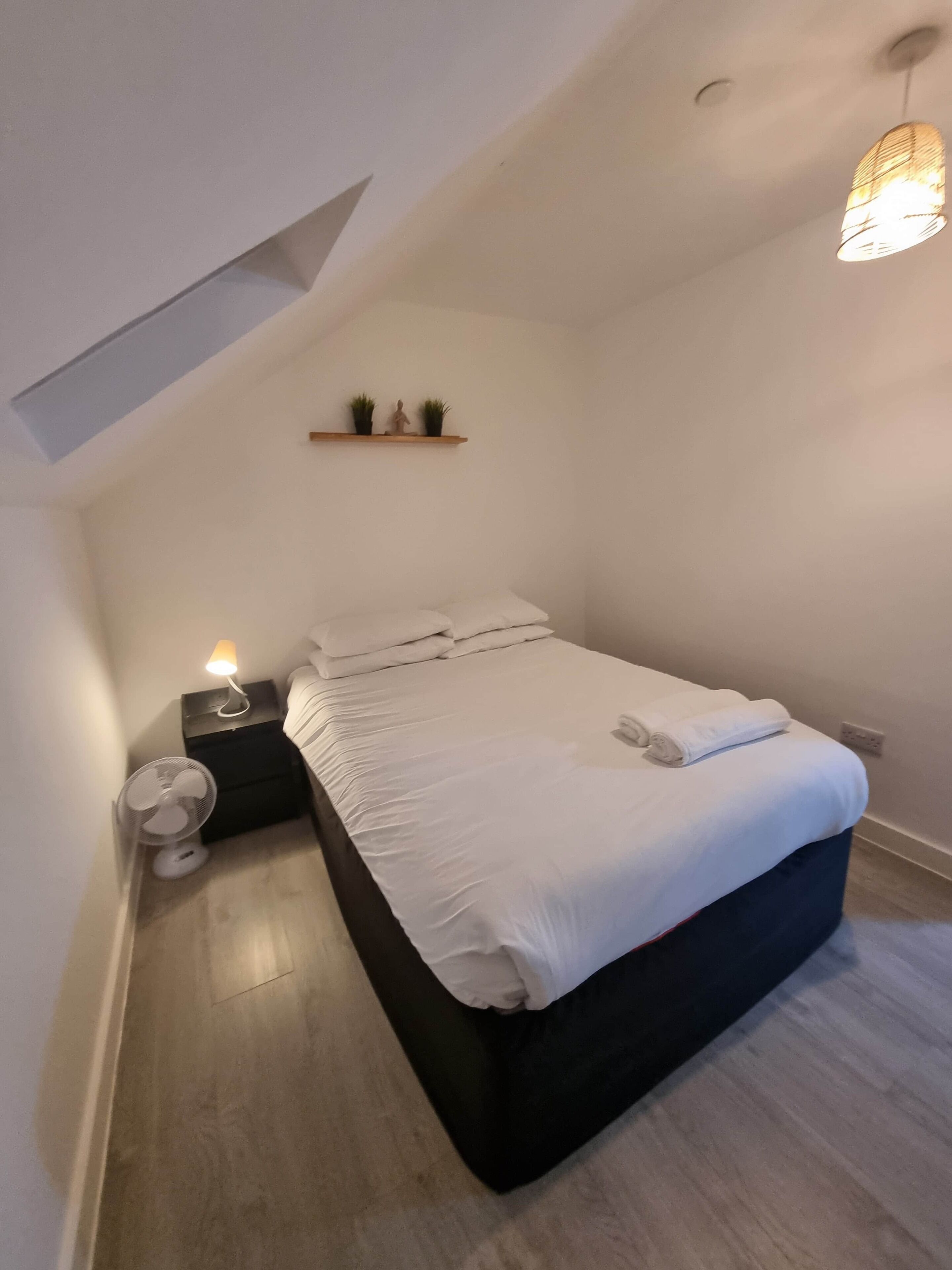 A modern bedroom with a bed, bedside table, lamp, and a small shelf with plants.