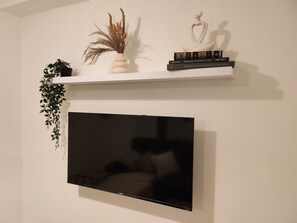 A flat-screen TV mounted on a wall, a white shelf with decorative items, and a hanging plant.