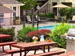 Eine Poolterrasse mit Holztischen und -bänken, umgeben von Grünpflanzen und einem Grill.