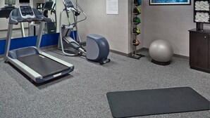Un gimnasio con cintas de correr, elípticas, una bicicleta estática, un televisor montado en la pared, un cartel motivacional, un estante con pelotas de ejercicios y una colchoneta para yoga.