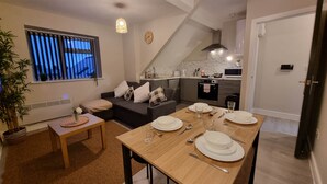 A modern living room with a dining table set for four, a sofa, a kitchen in the background, and a large window with blinds. - ApartHotel - Flat 7 (1 bed) (Cardiff)