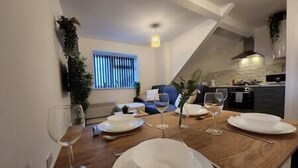 A dining area with a wooden table set for four, a kitchen in the background, and a window with blinds. - ApartHotel - Flat 7 (1 bed) (Cardiff)