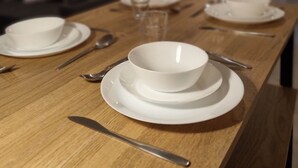 A dining table set with white plates, bowls, and cutlery, with a wooden table and a kitchen in the background. - ApartHotel - Flat 7 (1 bed) (Cardiff)