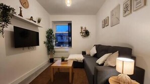 A living room with a flat-screen TV, a dark-colored sofa, a wooden coffee table, and a lamp on a side table. - ApartHotel - Flat 6 (1 bed) (Cardiff)