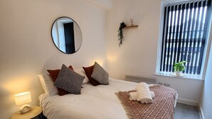 Une chambre bien rangée, avec un lit, une table de chevet, une lampe, un miroir et une fenêtre équipée de stores.