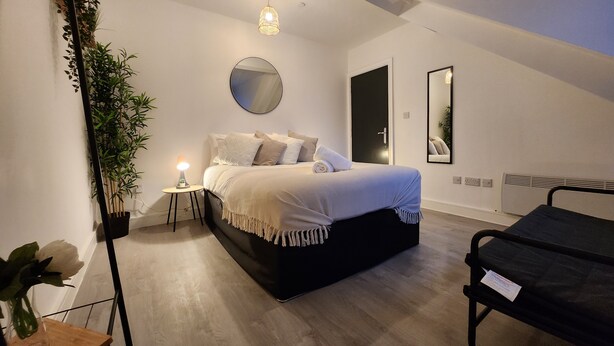 A bedroom with a bed, bedside table, lamp, mirror, and a potted plant.