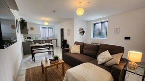A modern living room with a brown sofa, a coffee table, and a dining area with a bench. There is a kitchen in the background with a window and blinds. - ApartHotel - Flat 8 (2 bed) (Cardiff)