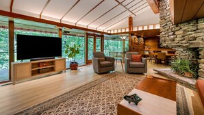 Amplia sala de estar con un televisor de pantalla plana grande, asientos cómodos, chimenea de piedra y ventanas amplias que ofrecen vistas al verdor.