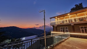 Un edificio de dos pisos con balcón, rodeado de barandilla metálica, con vistas a un paisaje montañoso al anochecer.