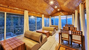 Interior de madera con asientos y mesas, ventanas amplias con cortinas y vistas a montañas.