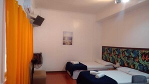 A hotel room with two beds, a small table, a chair, a fan, and a painting on the wall.