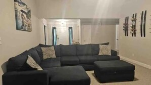 A living room with a sectional sofa, ottoman, and a painting on the wall.