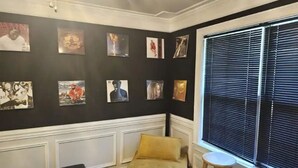 A room with a black wall adorned with framed artwork, a yellow sofa, a small stool, and a window with blinds.