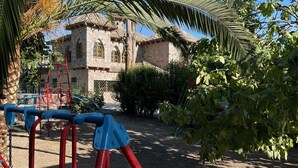 Ein Spielplatz mit roter und blauer Spielausstattung, einer Palme und einem Steinhaus im Hintergrund.