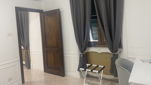 Una habitación con puerta de madera, ventana con cortinas oscuras, una mesita blanca y una silla gris.