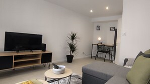 A modern living room with a flat-screen TV, a grey sofa, a small round table with a bowl and remote, a potted plant, and a dining area with a chair and table. - « Modern Cosy » Hyper Centre Bernay (Bernay)