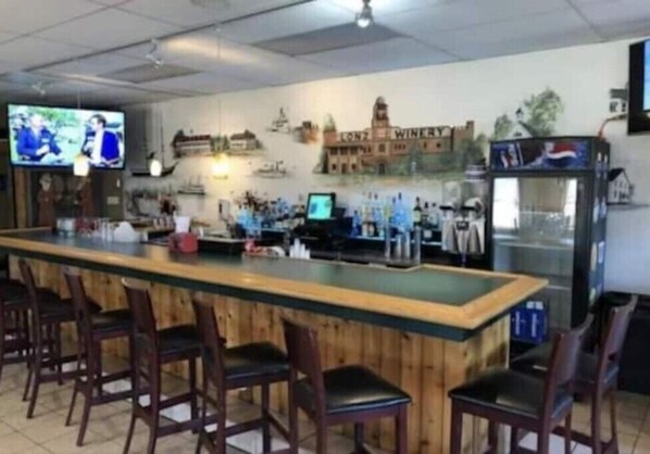 A bar area with a long counter, multiple bar stools, a television screen, and a wall mural depicting a building. - Middle Bass Island retreat,10-min ferry ride to Put-in-Bay, Pet friendly! (Middle Bass)