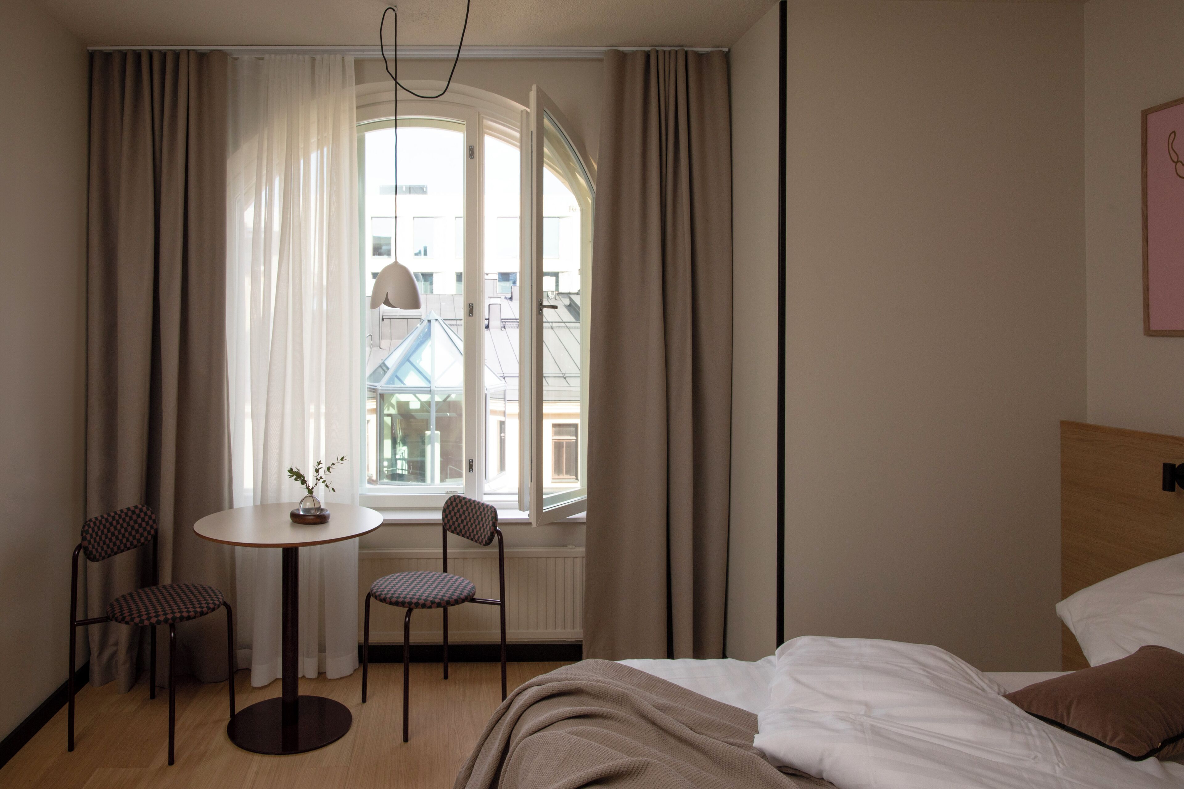 A hotel room with a bed, a round table with chairs, a view of buildings through a window, and a wall-mounted light fixture.