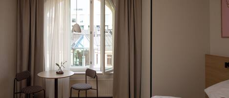 A hotel room with a bed, a round table with chairs, a view of buildings through a window, and a wall-mounted light fixture.