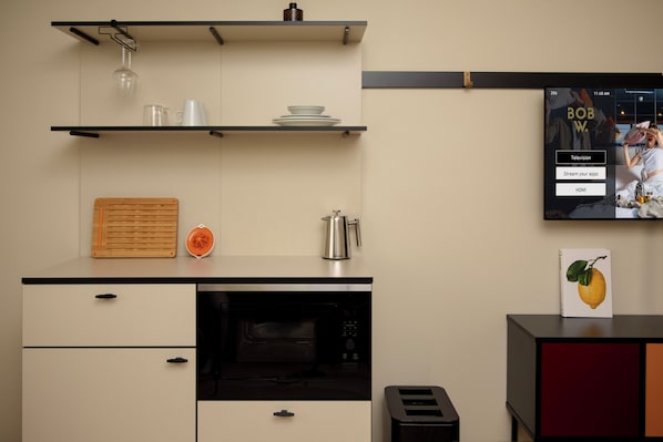 Una cocina moderna con microondas, tostadora y cafetera sobre la encimera. Hay estantes arriba con vajilla y un vaso.