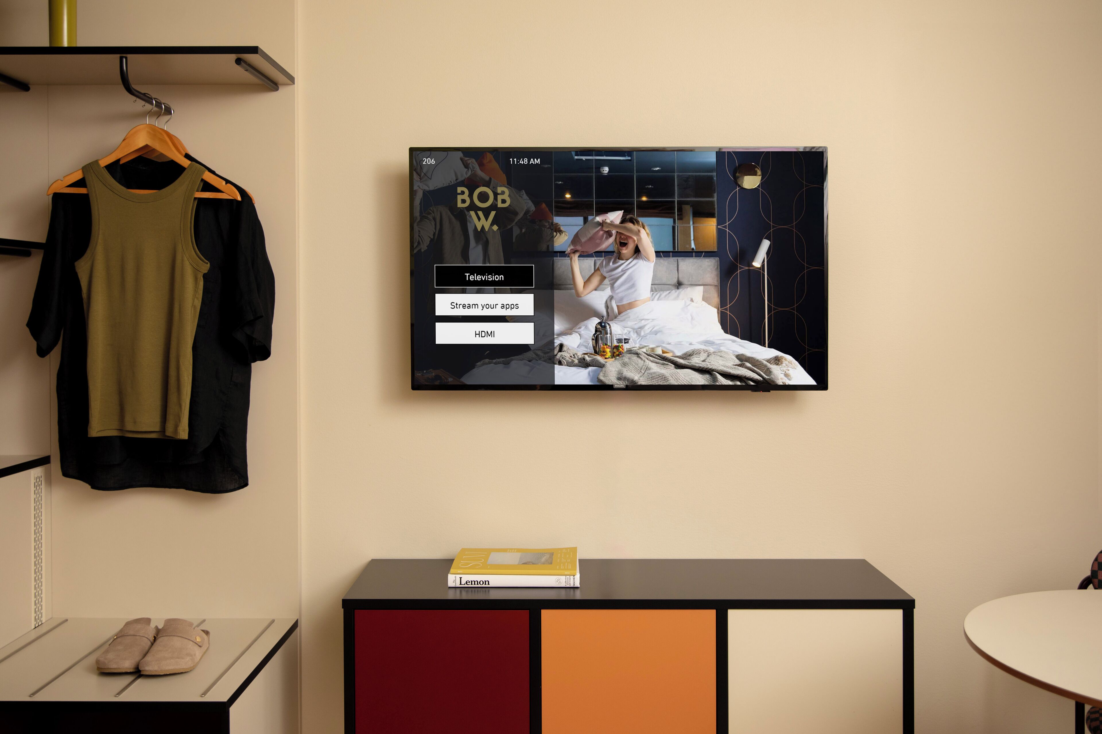 Una habitación con una televisión en la pared mostrando un programa, una cama con un almohadón, una mesita de noche con un libro y un perchero con una blusa verde y pantalones negros.