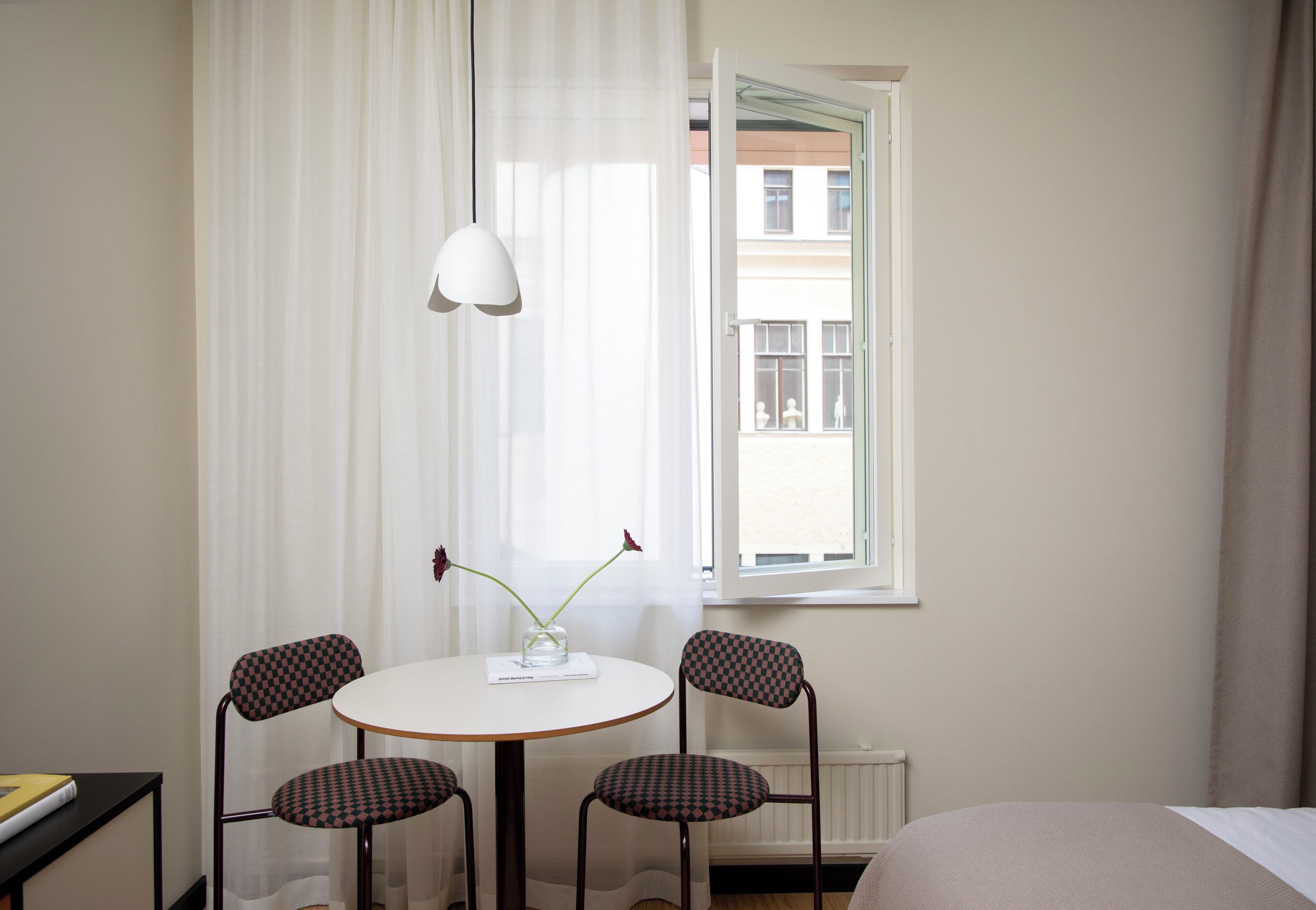 A hotel room with a round table, two checkered chairs, a vase with flowers, and a view of buildings through a window.