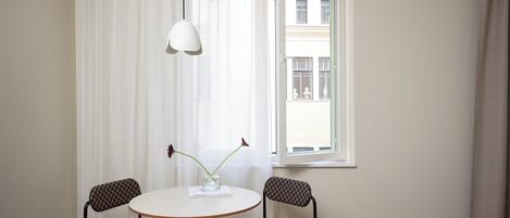 A hotel room with a round table, two checkered chairs, a vase with flowers, and a view of buildings through a window.