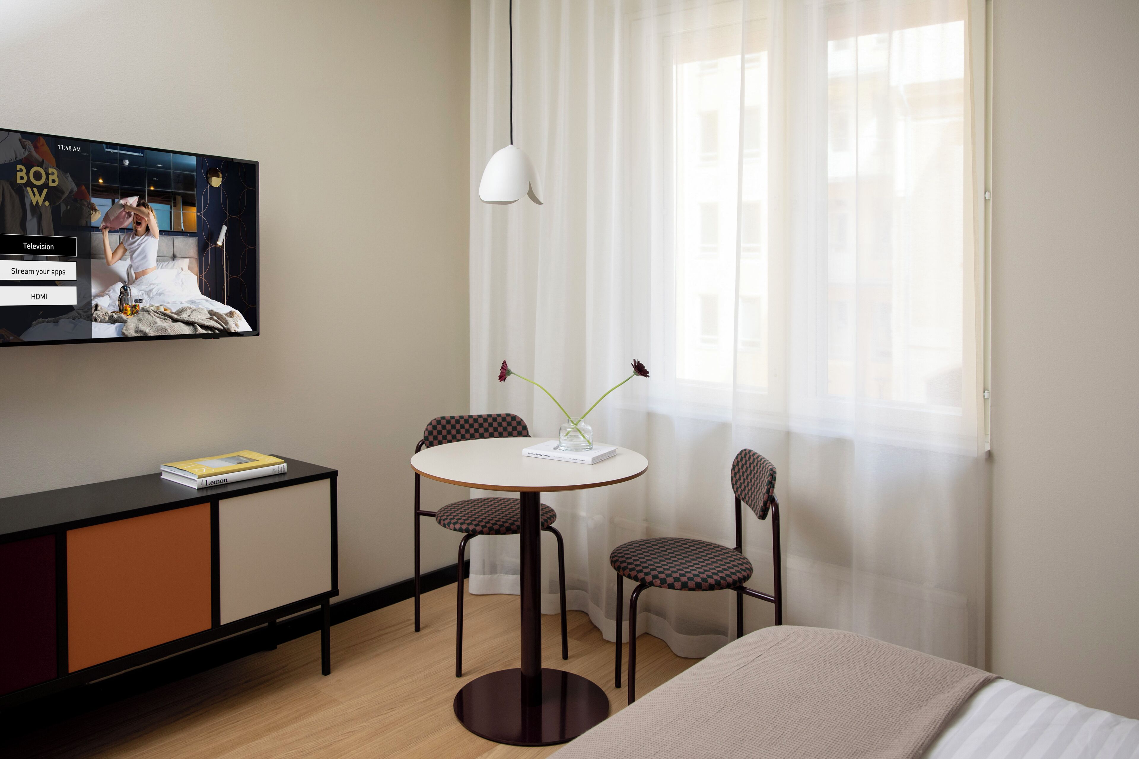 A hotel room with a bed, a TV mounted on the wall, a small round table with two chairs, a vase with flowers, and a window with sheer curtains.