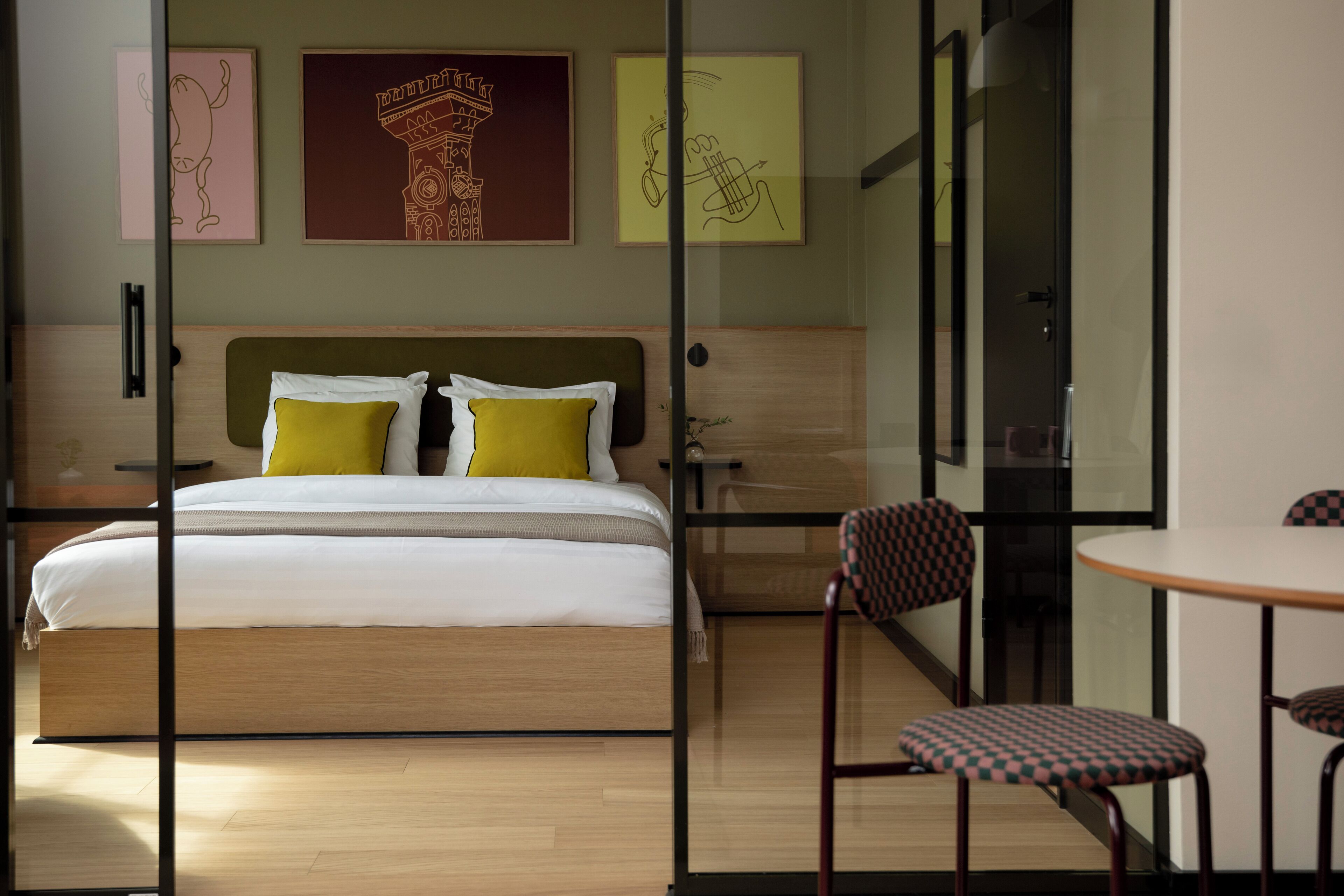 A modern hotel room with a wooden bed, two yellow pillows, a green headboard, and a glass partition. There is a small table with checkered chairs and a framed picture on the wall.