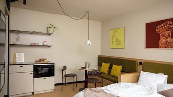 A compact hotel room with a kitchenette, a bed with white linens, a green upholstered headboard, and a small table with a chair.